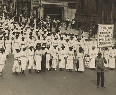 thousands of black women wearing white long dresses and some men in suits march in a 1917 New York City street. Three protest signs are visible.