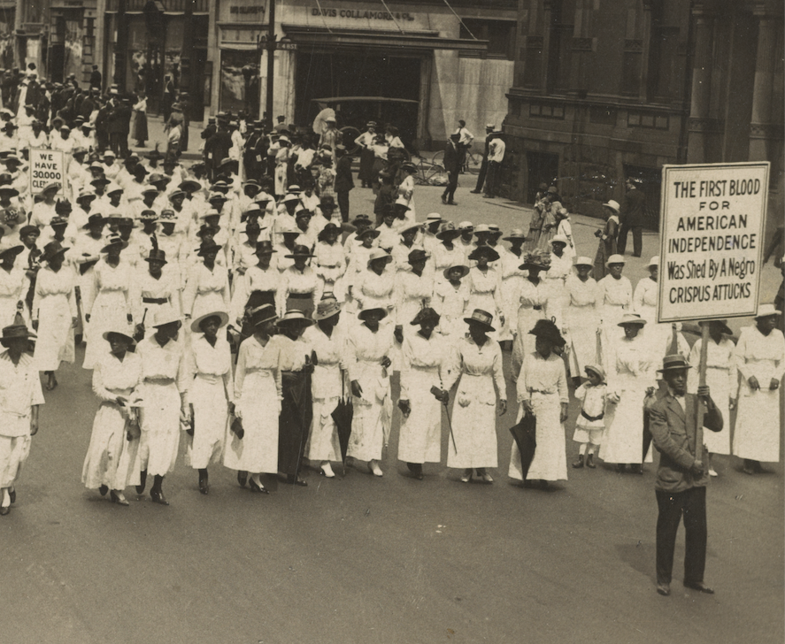 Rows of African American women wearing long white dresses, arms linked, fill a city street, led by a man holding a sign reading, "The First Blood for American Independence was Shed by a Negro: Crispus Atticus"