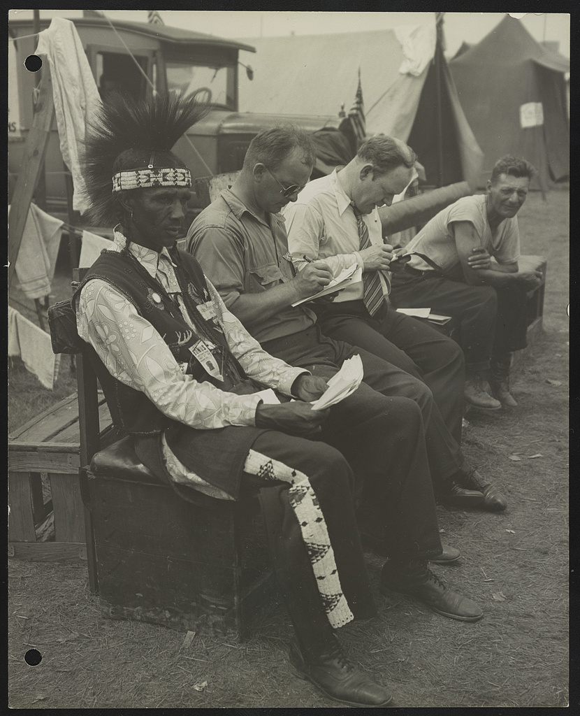 Photograph shows four men, probably veterans, sitting on a bench with tents in the background, probably at the Bonus Expeditionary Forces assemblage in Washington, D.C. One of the men is wearing a traditional Indian headdress, and pants and a vest with Indian motifs.