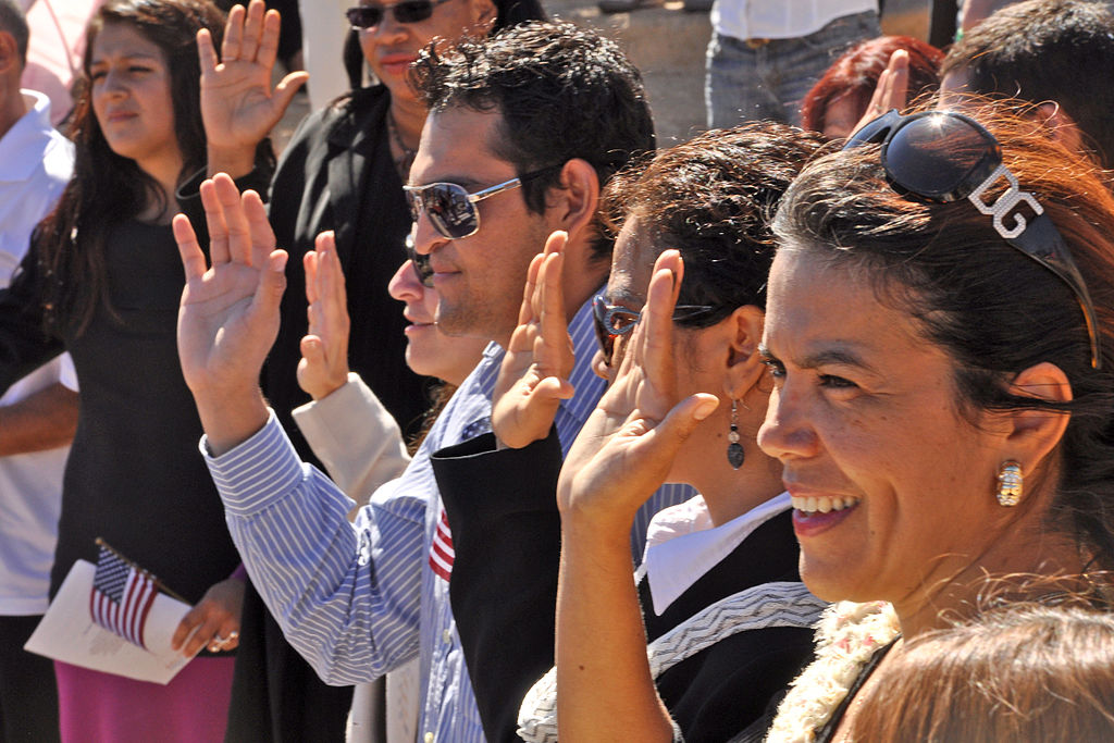 Naturalization ceremony close-up photo shows people with raised hands and smiles as they are sworn in as US citizens 