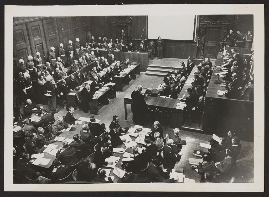 Photo of Nuremberg courtroom shows the many rows of judges, lawyers, helmeted military police, and defendants. 