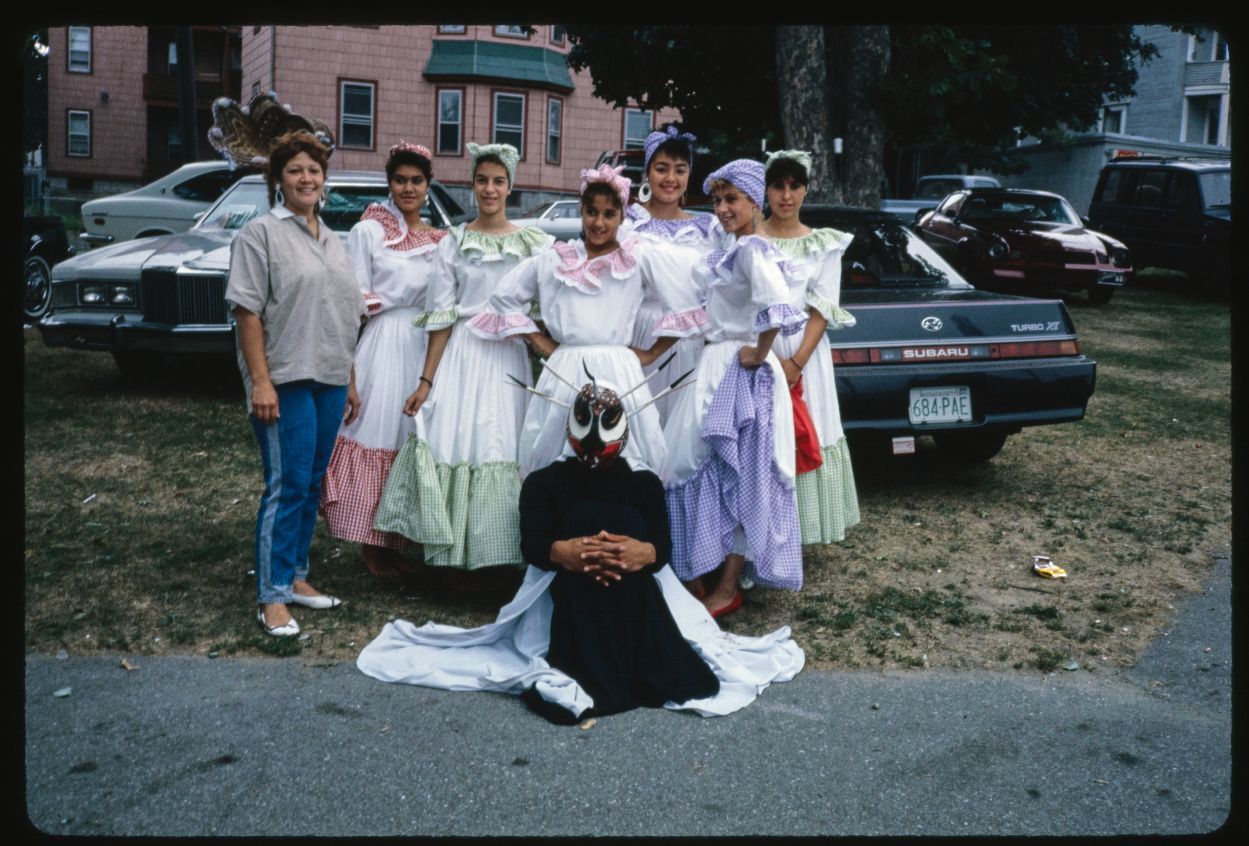 A group of smiling young women in long pastel dresses, white aprons, and kerchiefs on their heads pose outdoors with a black masked and costumed figure at their feet, and an older woman in jeans and casual shirt beside them. In the background is a house and parked cars. 