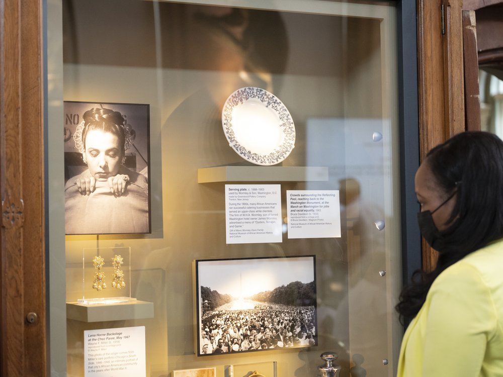 Photographs and objects are mounted on a wall in a glass display case 