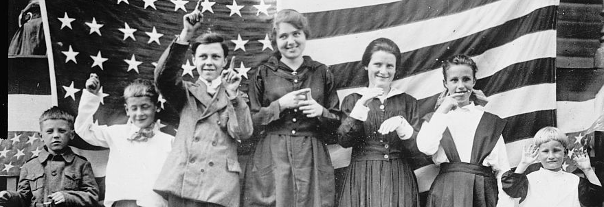 Young students smile and sign the Star Spangled Banner, in front of 48-star United States flag.