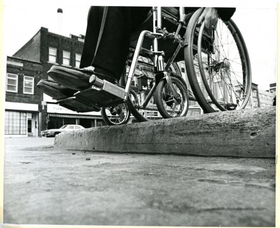 Inaccessible curb, mid-20th century. A wheelchair is rolled right up to the edge. 