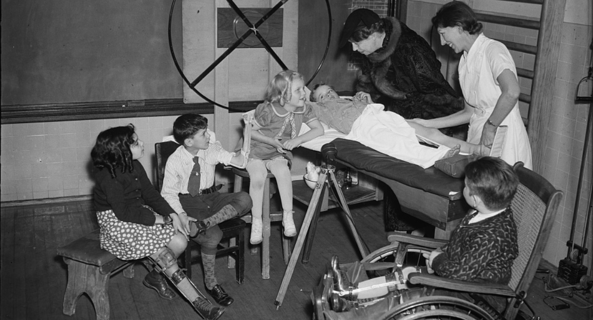 A classroom in a children's school with a blackboard and equipment in the background. A group of children are sitting or standing in front of a medical table, and one boy is laying down in front of the table. A nurse in a white uniform leans over the boy, adjusting his leg. Eleanor Roosevelt, wearing a large fur coat, smiles down at the boy from behind the table. The children have cheerful expressions. 