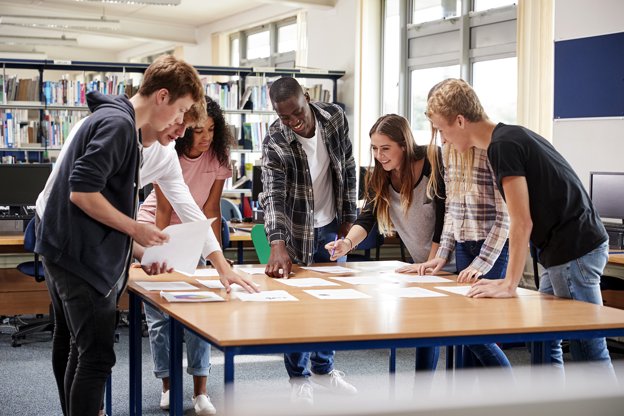 A diverse group of seven high school students lean over to study several documents laid out on a table in the library. 