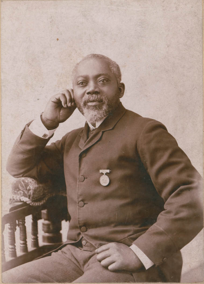 An older Black man, balding with a white beard. He wears a medal on his dress coat. He poses seated, with one hand aside his cheek and the other on his thigh. He looks past the camera. 