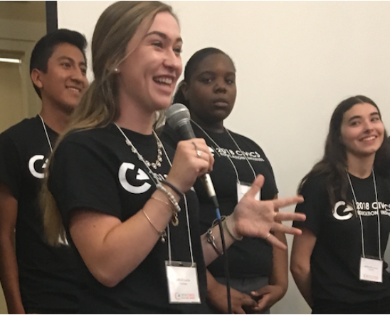 A diverse group of high school students presents on civic engagement at a state educators institute. The speaker has long hair, she is smiling as she holds a microphone. All the students are wearing black civic engagement t-shirts.