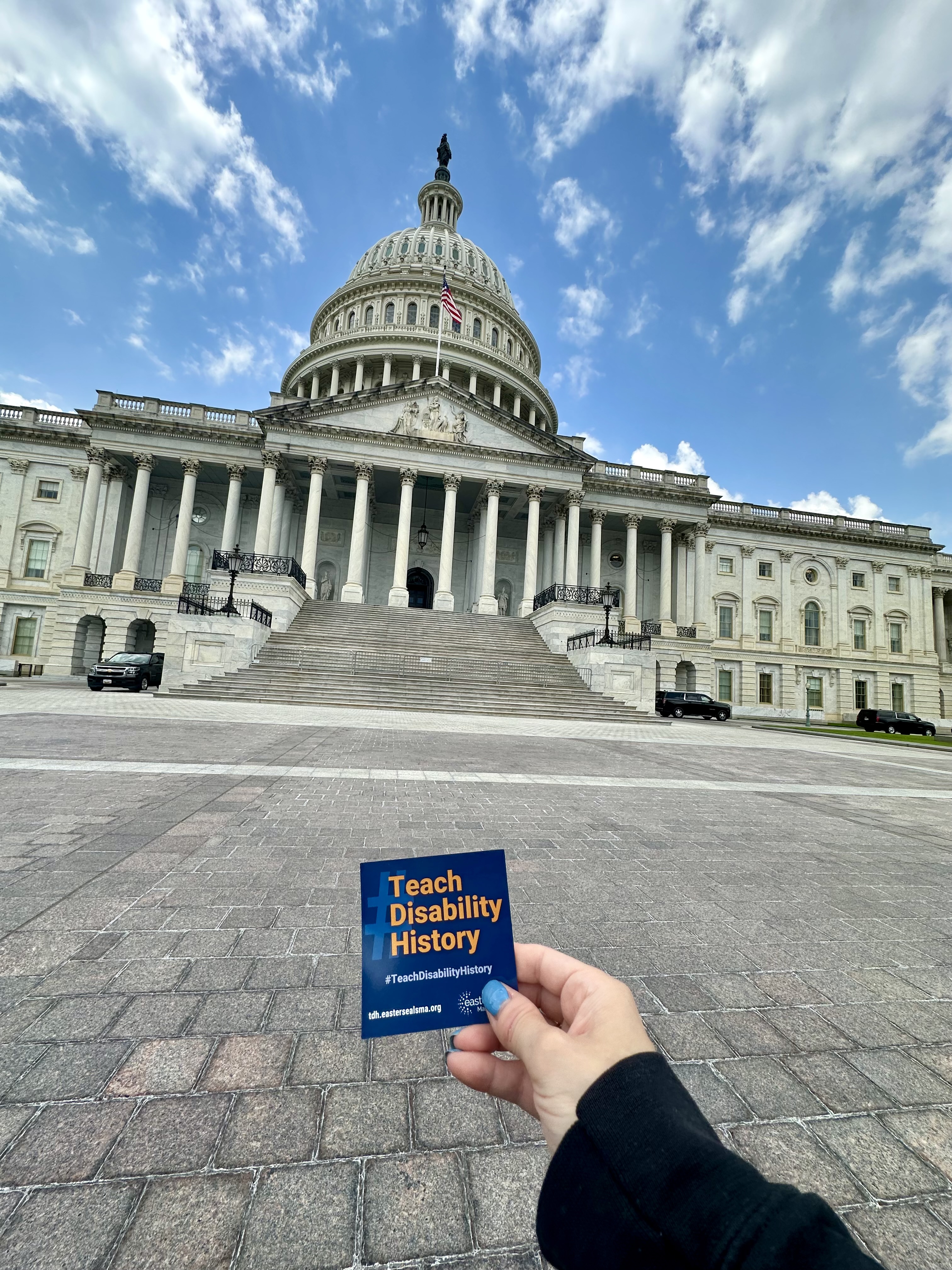 A hand holds a card reading "Teach Disability History" in front of the steps of the U.S. Capitol site of the 1990 Capitol Crawl protest