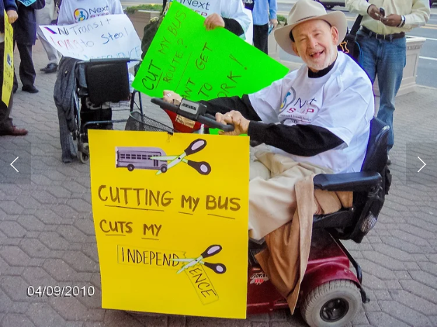 On the street during a march, an man in a cowboy hat and t-short using a wheelchair holds a sign reading "Cutting my bus cuts my independence" 
