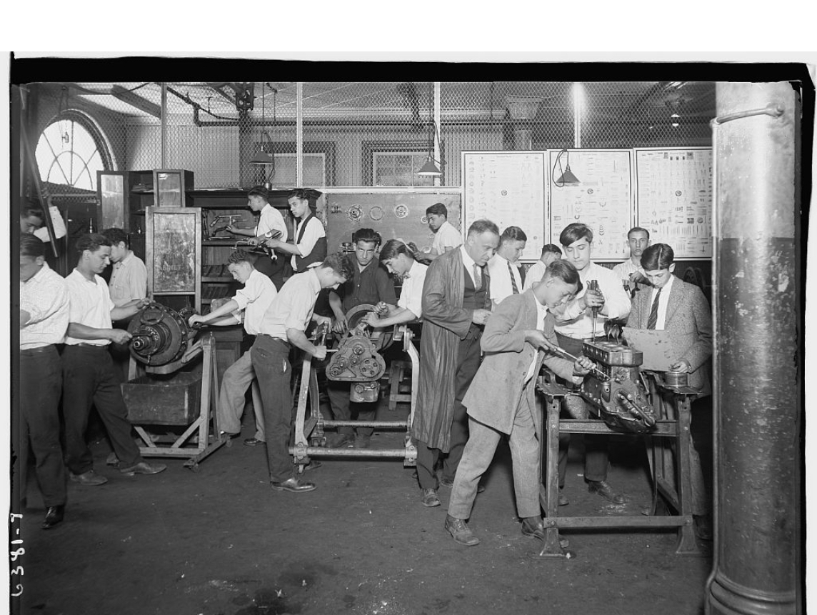 Youths work in a machine shop supervised by a man in a long coat.