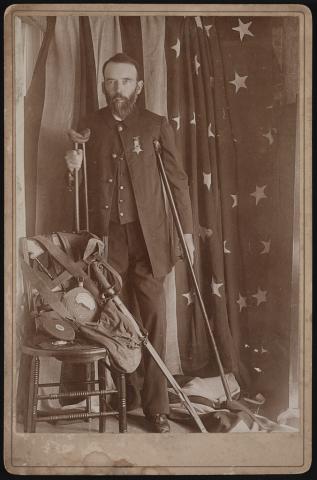 Civil War veteran and amputee Henry A. Seaverns of G.A.R. George W. Perry Post no. 31, Scituate, Massachusetts, in uniform with sword, canteen, and other artifacts, standing on crutches in front of American flag.