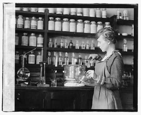 In front of shelves of labeled bottles and a lab bench with flasks, a young woman extracts something with tongs.