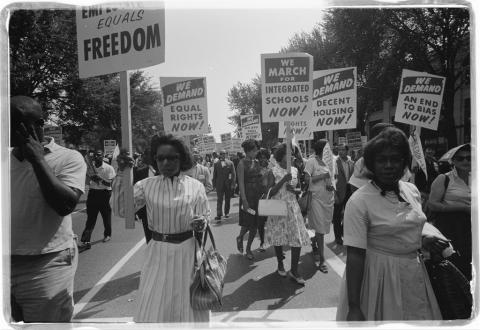 Black Americans peacefully walk in the center of the street with protest signs demanding freedom, equality, integrated schools, the women in summer dresses with handbags.