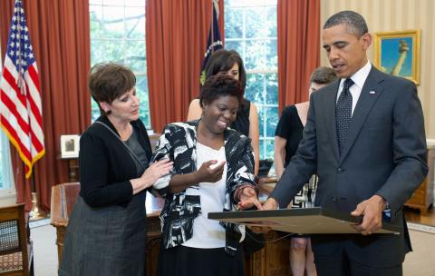Woman with short Afro touches her framed artwork held by the president, who wears a suit. A second woman holds the artist's arm while looking at the picture. 