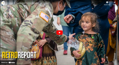 A uniformed and masked U.S. soldier bends to touch the shoulder of a young blonde girl carrying two bananas in a plastic bag and smiling