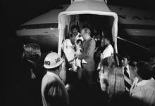 President Ford cradles a baby on the steps to a jet plane with guards and staff around