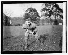 A young man in a sweatshirt and sweat pants leans forward as he hugs a football