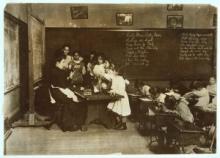 A classroom with students working at desks, teacher in long dress seated at a table next to chalk boards, surrounded by a group of 6 girls of varied heights, using her hands as she explains something.