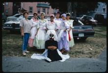 Several young women pose in traditional dresses. A person in a bird costume sits on the ground. 