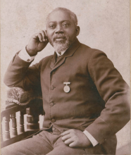 African American William H. Carney, balding and with a trimmed white beard, sits, wearing a civilian suit, and with a medal pinned to his chest.