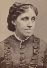 Posed photo of a middle-aged woman with hair bound behind her head and wearing a dress with ruffles up both sides of the front and over her shoulders. She wears white lace at the neck and a white cross. She looks to her left beyond the camera. 