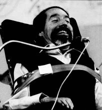 Ed Roberts, a smiling man with dark hair and beard is strapped into his wheelchair. He is speaking into a microphone.
