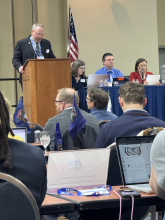 Casey Cullen, a white man in a suit, leads a meeting from a podium. There is an American flag behind him. Two women and a man sit at the table to his left. Several delegates sit facing front with their laptops open. 