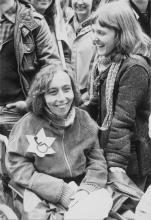 A young woman using a wheelchair and another young woman companion laugh. The first woman wears a large Star of David with a handicapped symbol of a wheelchair on it. Both women are white, have shoulder-length light brown hair and wear heavy coats. They are part of a larger group also wearing warm clothes.