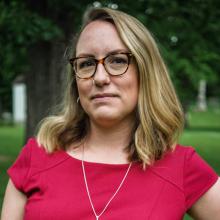 A blond woman with shoulder length hair and glasses stands on the leafy green of a campus. She wears glasses, a short-sleeved shirt and a delicate gold necklace.
