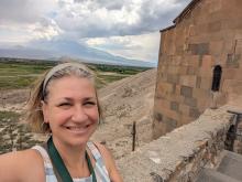 A smiling white woman with short hair takes a selfie as she stands atop an ancient stone building with a dry landscape of farms and mountains behind her. 