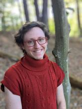 Outdoors in the woords, a smiling Max Chervin Bridge wears short hair, glasses and a turtleneck sweater