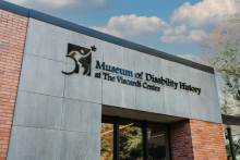 Exterior of the Museum of Disability History at The Viscardi Center shows the glass doorway. The museum logo is a person using a wheelchair holding aloft a star. 