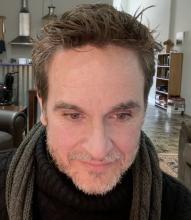 A white man with short brown hair and a gray and brown beard sits in a marble-floored living room. He wears a turtle-neck sweater and has a knit scarf draped around his neck. A wheelchair is in the background.
