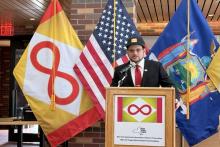 A man in a suit and red tie speaks at a podium indoors, with a microphone in front of him. Behind him are three flags: the U.S. flag, the New York State flag, and a red, yellow, and white flag that is the New York State Neurodiversity Strength Flag.