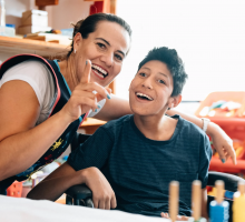 A smiling teacher and student, both with straight dark hair in a classroom - her arm is on the back of his wheelchair - his head is cocked at an angle