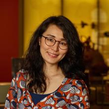 A smiling Asian-American woman with long dark hair and glasses wears a colorful shirt with a bird print. 