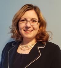 A white woman with shoulder length hair, and wearing glasses a gold necklace and a blazer smiles for the camera