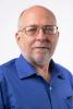 "A balding white man with glasses and a white beard, wearing a button shirt with no tie.